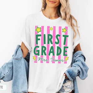 May include: A white short-sleeved t-shirt featuring a graphic with vertical pink stripes and the green text FIRST GRADE. Below the text is the name Mrs. Tuner in green cursive script, accented by yellow star and lightning bolt illustrations.