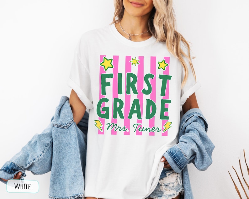 May include: A white short-sleeved t-shirt featuring a graphic with vertical pink stripes and the green text FIRST GRADE. Below the text is the name Mrs. Tuner in green cursive script, accented by yellow star and lightning bolt illustrations.