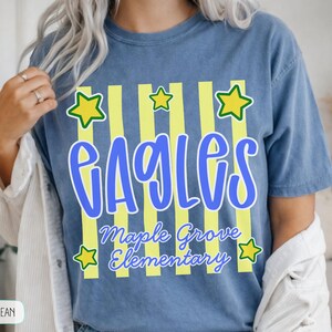 May include: A blue jean colored t-shirt featuring a graphic with vertical yellow stripes and yellow stars. The text reads EAGLES in large blue letters, with Maple Grove Elementary written below in a smaller blue script font.