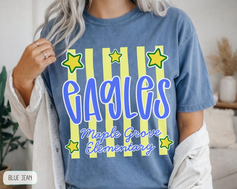 May include: A blue jean colored t-shirt featuring a graphic with vertical yellow stripes and yellow stars. The text reads EAGLES in large blue letters, with Maple Grove Elementary written below in a smaller blue script font.