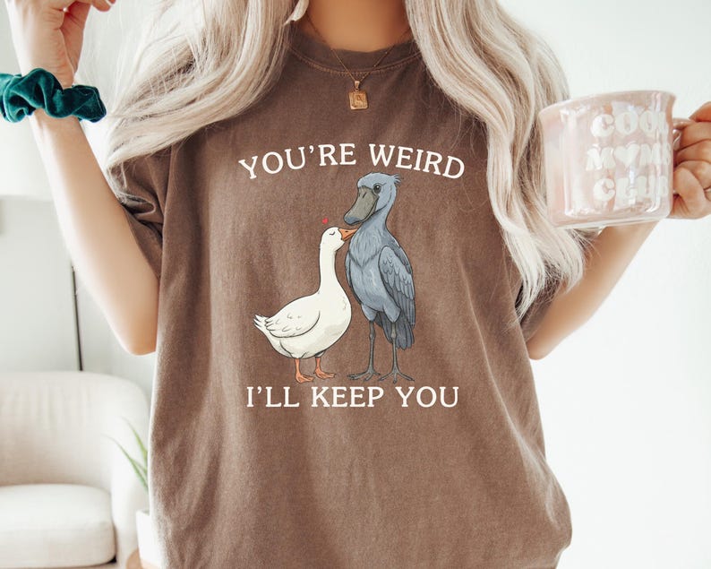 May include: Brown t-shirt with the text "YOU'RE WEIRD I'LL KEEP YOU" and a graphic of a white goose and a gray shoebill bird. The goose and bird are facing each other. The person is holding a pink mug.