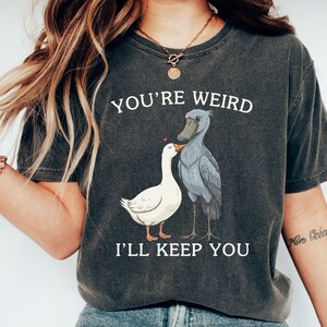 May include: Dark gray t-shirt with the text "YOU'RE WEIRD I'LL KEEP YOU" above an illustration of a white goose and a gray shoebill stork. The goose has a red heart above its head. The shirt is a casual style.