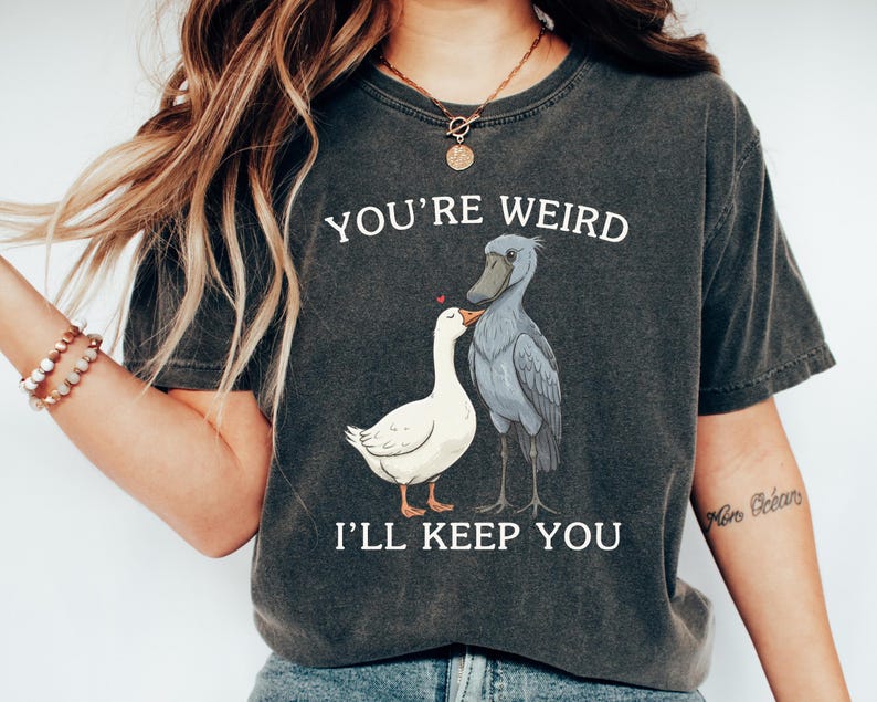 May include: Dark gray t-shirt with the text "YOU'RE WEIRD I'LL KEEP YOU" above an illustration of a white goose and a gray shoebill stork. The goose has a red heart above its head. The shirt is a casual style.