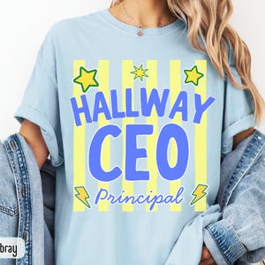 May include: A light blue t-shirt featuring a graphic with vertical yellow stripes, green stars, and yellow lightning bolts. The text reads "HALLWAY CEO" in large blue letters and "Principal" in smaller blue script font across the center.