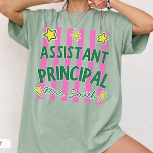 May include: A sage green short-sleeved t-shirt featuring a graphic with vertical pink stripes. The text reads ASSISTANT PRINCIPAL and Mrs. Smith in green, accented by yellow stars and lightning bolts. The shirt has a crew neckline and loose fit.