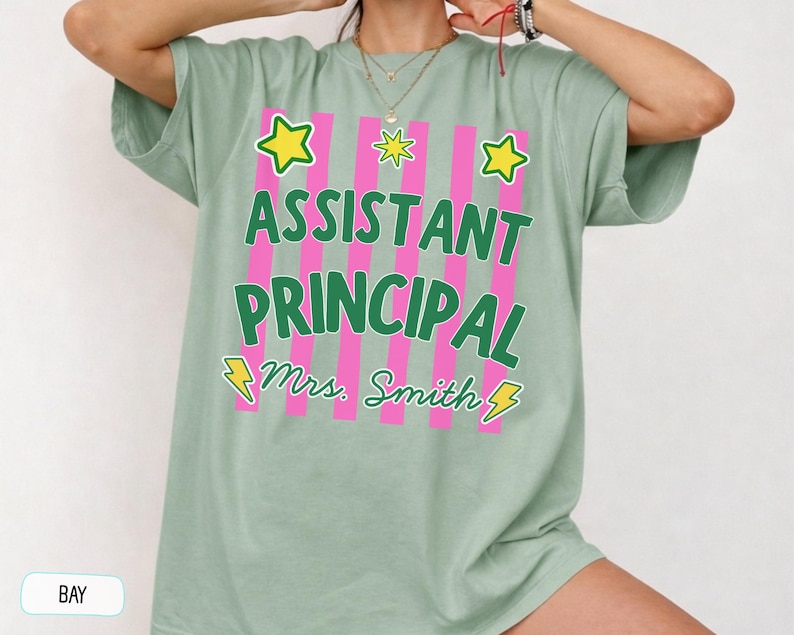 May include: A sage green short-sleeved t-shirt featuring a graphic with vertical pink stripes. The text reads ASSISTANT PRINCIPAL and Mrs. Smith in green, accented by yellow stars and lightning bolts. The shirt has a crew neckline and loose fit.
