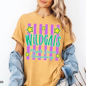 May include: A mustard yellow short-sleeve t-shirt featuring a graphic with vertical purple stripes, yellow stars, and green text that reads WILDCATS Maple Grove Elementary. The shirt has a crew neckline and a relaxed fit with rolled sleeves.