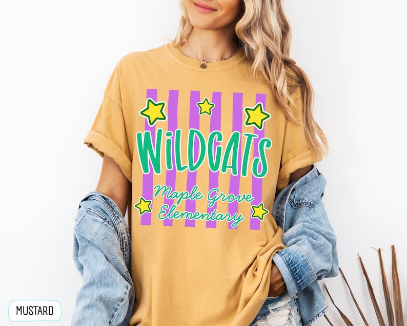 May include: A mustard yellow short-sleeve t-shirt featuring a graphic with vertical purple stripes, yellow stars, and green text that reads WILDCATS Maple Grove Elementary. The shirt has a crew neckline and a relaxed fit with rolled sleeves.