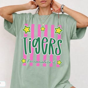 May include: A sage green short-sleeve t-shirt featuring a graphic design with vertical pink stripes, the word "Tigers" in large green script, the text "Personalize it" in smaller green script, and five yellow stars with green outlines.