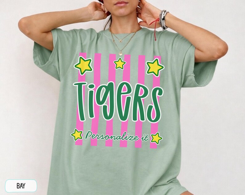 May include: A sage green short-sleeve t-shirt featuring a graphic design with vertical pink stripes, the word "Tigers" in large green script, the text "Personalize it" in smaller green script, and five yellow stars with green outlines.