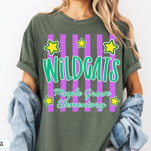May include: A moss green t-shirt featuring a graphic with vertical purple stripes, yellow stars, and the text "WILDCATS" in large green letters above "Maple Grove Elementary" in smaller green script. The shirt has a crew neckline and short sleeves.