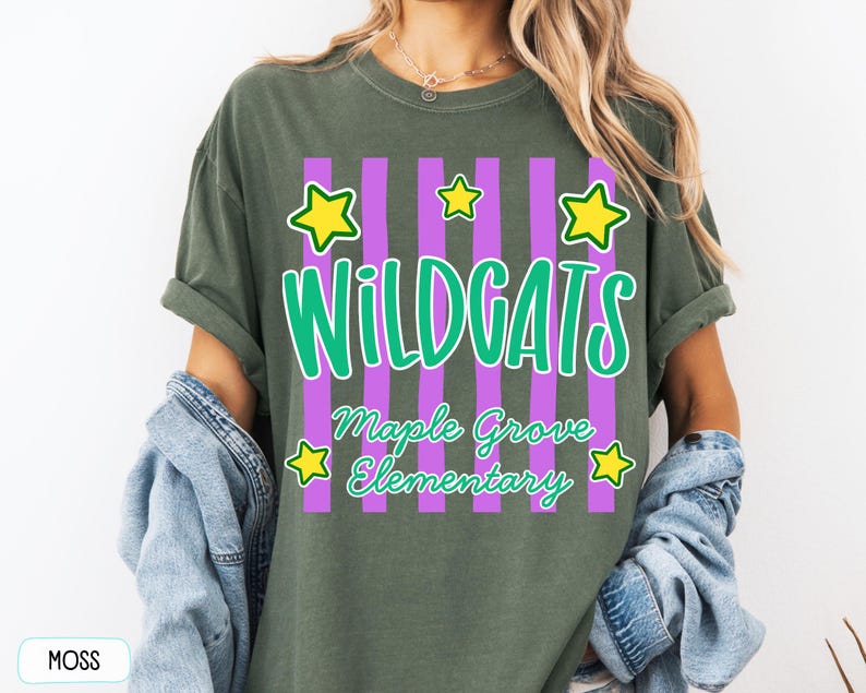 May include: A moss green t-shirt featuring a graphic with vertical purple stripes, yellow stars, and the text "WILDCATS" in large green letters above "Maple Grove Elementary" in smaller green script. The shirt has a crew neckline and short sleeves.