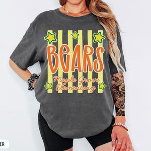 May include: A charcoal grey short-sleeve t-shirt featuring a graphic design with vertical yellow stripes, orange text that reads "BEARS", and smaller orange text that reads "Maple Grove Elementary". The design includes five yellow stars.