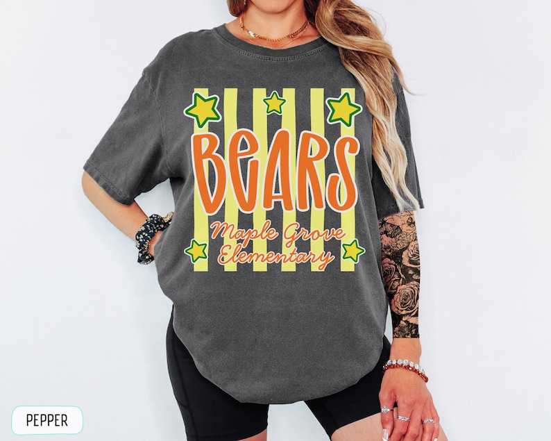 May include: A charcoal grey short-sleeve t-shirt featuring a graphic design with vertical yellow stripes, orange text that reads "BEARS", and smaller orange text that reads "Maple Grove Elementary". The design includes five yellow stars.