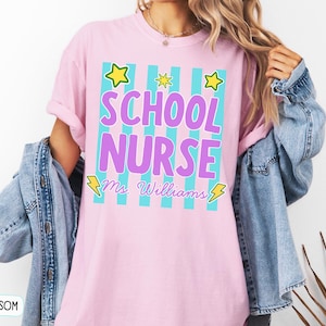 May include: A light pink short-sleeve t-shirt featuring a graphic with vertical teal stripes, yellow stars, and yellow lightning bolts. The text reads "SCHOOL NURSE" in large purple letters, with "Ms. Williams" written below in a cursive script.