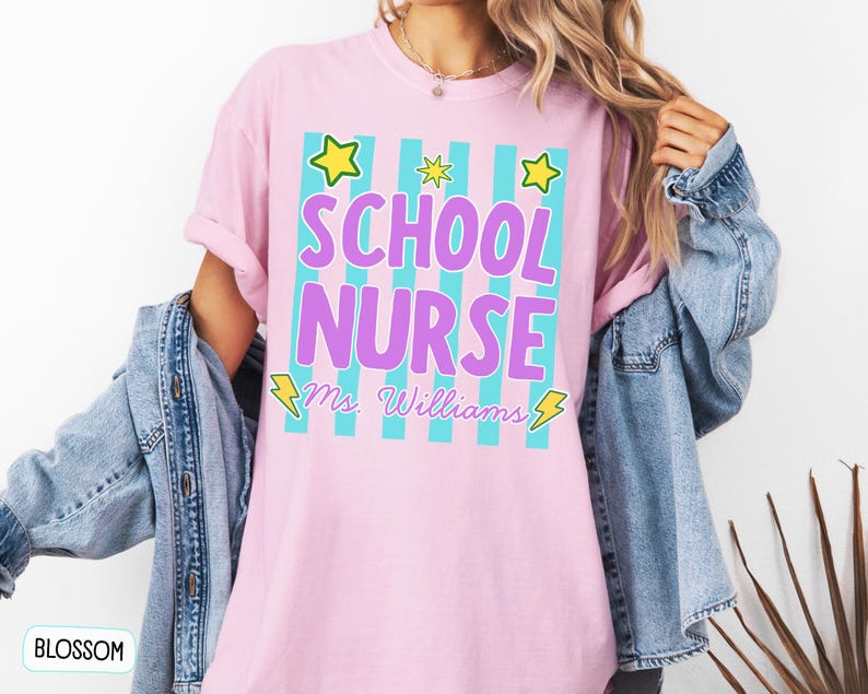 May include: A light pink short-sleeve t-shirt featuring a graphic with vertical teal stripes, yellow stars, and yellow lightning bolts. The text reads "SCHOOL NURSE" in large purple letters, with "Ms. Williams" written below in a cursive script.