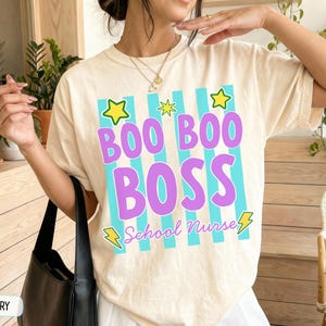 May include: An ivory t-shirt featuring a graphic design with vertical cyan stripes, yellow stars, and yellow lightning bolts. The text reads BOO BOO BOSS in large purple letters, with the words School Nurse in smaller purple cursive script below.