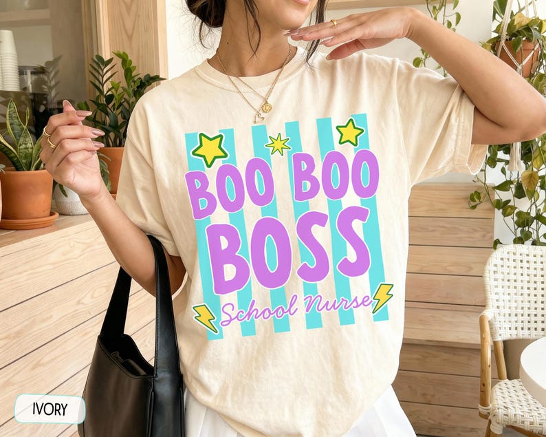 May include: An ivory t-shirt featuring a graphic design with vertical cyan stripes, yellow stars, and yellow lightning bolts. The text reads BOO BOO BOSS in large purple letters, with the words School Nurse in smaller purple cursive script below.