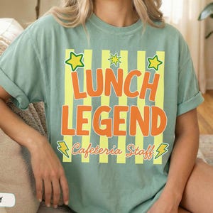 May include: A sage green short-sleeve t-shirt featuring a graphic print with vertical yellow stripes. The text reads LUNCH LEGEND in large orange letters, with Cafeteria Staff in cursive below. Yellow stars and lightning bolts accent the design.