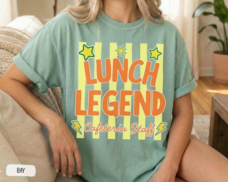 May include: A sage green short-sleeve t-shirt featuring a graphic print with vertical yellow stripes. The text reads LUNCH LEGEND in large orange letters, with Cafeteria Staff in cursive below. Yellow stars and lightning bolts accent the design.