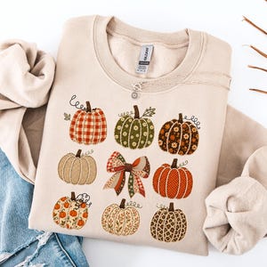 May include: A beige crewneck sweatshirt featuring a graphic print of eight pumpkins and a central bow. The pumpkins display various patterns including gingham, floral, and polka dots in shades of orange, green, and brown. A silver necklace rests on top.
