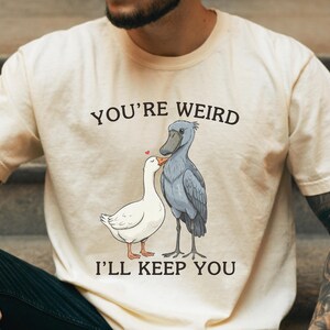 May include: Cream-colored t-shirt with the text "YOU'RE WEIRD I'LL KEEP YOU" above a cartoon illustration of a white goose and a gray Shoebill stork. The goose has a red heart above its head.