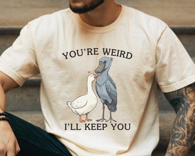 May include: Cream-colored t-shirt with the text "YOU'RE WEIRD I'LL KEEP YOU" above a cartoon illustration of a white goose and a gray Shoebill stork. The goose has a red heart above its head.