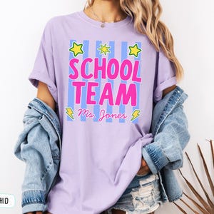 May include: An orchid-colored t-shirt featuring a graphic with vertical light blue stripes, three yellow stars, and two yellow lightning bolts. The text reads "SCHOOL TEAM" in large pink letters, with "Ms. Jones" written in cursive pink script below.
