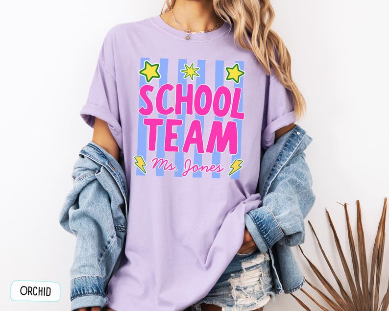 May include: An orchid-colored t-shirt featuring a graphic with vertical light blue stripes, three yellow stars, and two yellow lightning bolts. The text reads "SCHOOL TEAM" in large pink letters, with "Ms. Jones" written in cursive pink script below.