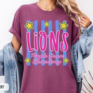 May include: A berry-colored t-shirt featuring a graphic with vertical light blue stripes, five yellow stars with green outlines, and the text "LIONS" in large pink letters above "Maple Grove Elementary" in smaller pink cursive script lettering.
