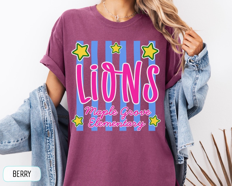 May include: A berry-colored t-shirt featuring a graphic with vertical light blue stripes, five yellow stars with green outlines, and the text "LIONS" in large pink letters above "Maple Grove Elementary" in smaller pink cursive script lettering.