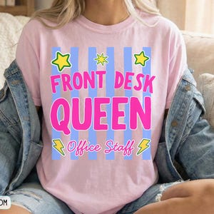 May include: A light pink t-shirt featuring a graphic with vertical blue stripes, yellow stars, and yellow lightning bolts. The text reads FRONT DESK QUEEN in large pink letters, with the words Office Staff in smaller pink cursive script below.