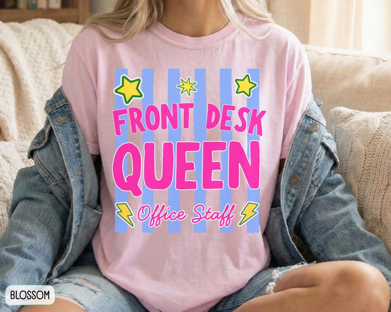 May include: A light pink t-shirt featuring a graphic with vertical blue stripes, yellow stars, and yellow lightning bolts. The text reads FRONT DESK QUEEN in large pink letters, with the words Office Staff in smaller pink cursive script below.