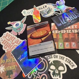 May include: A collection of holographic stickers and a Magic: The Gathering card on a black surface. Stickers include a frog, a skull, a hot sauce bottle, and text that reads "Lizards of the Coast" and "Clue".