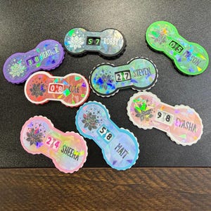 May include: A collection of colorful, personalized name tags with a unique design. Each tag features a name and a number, with a floral design and holographic accents. The tags are in various colors, including purple, green, and pink.