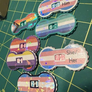 Pu&ograve; includere: Una collezione di dadi con pronomi colorati con vari disegni di bandiere del pride. Ogni dado presenta un numero e i pronomi "He/Him" o "She/Her". I dadi sono posizionati su uno sfondo a griglia verde.