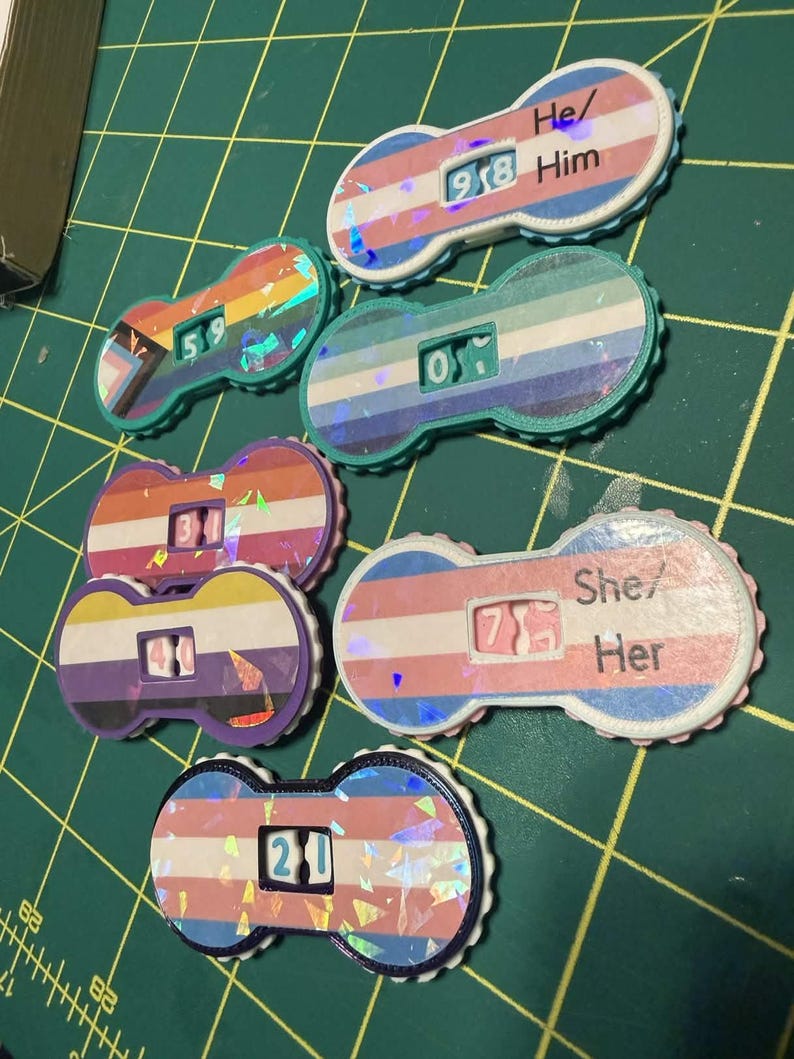 Pu&ograve; includere: Una collezione di dadi con pronomi colorati con vari disegni di bandiere del pride. Ogni dado presenta un numero e i pronomi "He/Him" o "She/Her". I dadi sono posizionati su uno sfondo a griglia verde.