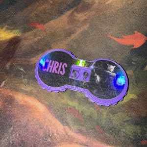 Pu&ograve; includere: Un accessorio viola a forma di chiave con il nome "CHRIS" in testo bianco. L'accessorio ha un centro trasparente con una grafica e i numeri "5 9". I bordi sono smerlati.