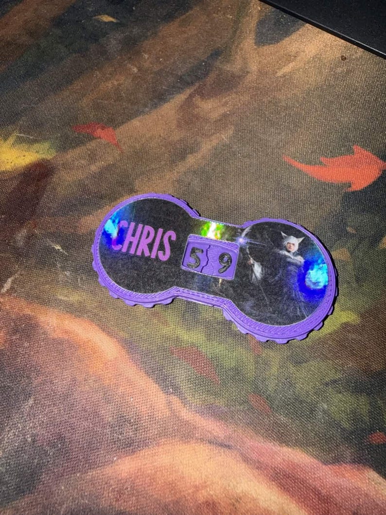 Pu&ograve; includere: Un accessorio viola a forma di chiave con il nome "CHRIS" in testo bianco. L'accessorio ha un centro trasparente con una grafica e i numeri "5 9". I bordi sono smerlati.