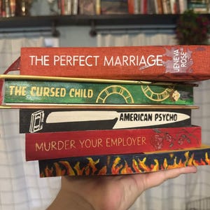 May include: A stack of books with titles including "The Perfect Marriage," "The Cursed Child," "American Psycho," and "Murder Your Employer." The books are held in a hand, with a bookshelf in the background.
