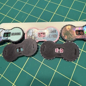 Pu&ograve; includere: Una collezione di quadranti contatore neri e colorati con vari disegni e testo, tra cui "Brianna" e "Other/She/Her". I quadranti hanno un display digitale che mostra i numeri. Lo sfondo &egrave; un tappetino da taglio verde.