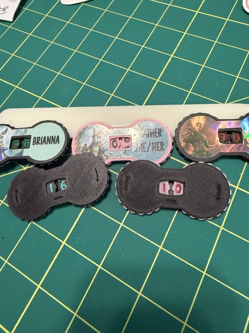 Pu&ograve; includere: Una collezione di quadranti contatore neri e colorati con vari disegni e testo, tra cui "Brianna" e "Other/She/Her". I quadranti hanno un display digitale che mostra i numeri. Lo sfondo &egrave; un tappetino da taglio verde.