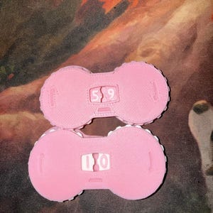 Pu&ograve; includere: Due oggetti rosa a forma di manubrio con numeri bianchi al centro. L'oggetto superiore mostra il numero 59, e quello inferiore 10. Gli oggetti hanno una superficie strutturata e un bordo a pois.