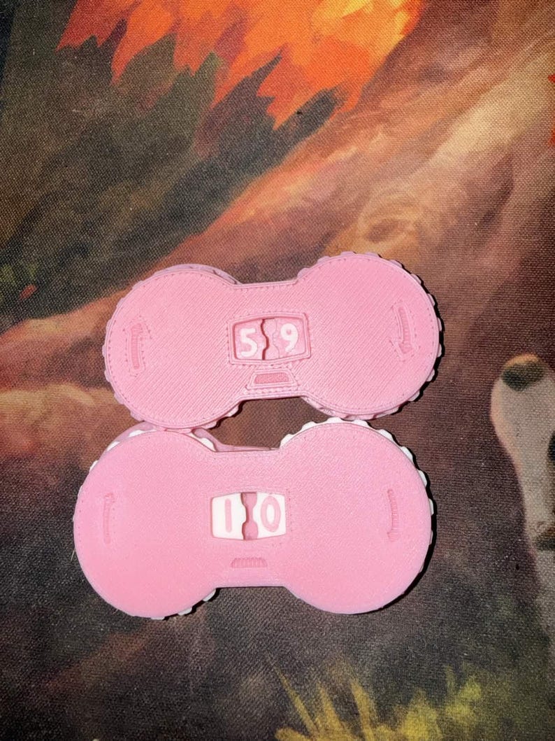 Pu&ograve; includere: Due oggetti rosa a forma di manubrio con numeri bianchi al centro. L'oggetto superiore mostra il numero 59, e quello inferiore 10. Gli oggetti hanno una superficie strutturata e un bordo a pois.