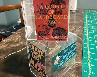 Può includere: Una teca in acrilico trasparente con una carta rossa che riporta il testo A COURT OF CARDBOARD CRACK MAGIC T. GATHERING. Il lato della teca mostra un adesivo con il testo GRAVE CRAWLER MAGIC THE GATHERING e 50% MTG 50% BOOKS 100% READING.