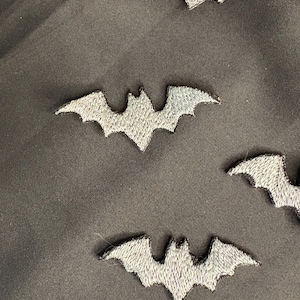 Può includere: Primo piano di diverse toppe a forma di pipistrello ricamate in grigio su uno sfondo di tessuto nero. Le toppe a forma di pipistrello presentano dettagliati disegni di ali e corpo, perfetti per l'artigianato a tema Halloween o gotico.