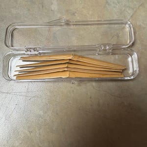 May include: A clear plastic case, open, containing several thin, light brown reeds. The reeds are stacked neatly inside the rectangular case. The case is on a neutral-toned surface.