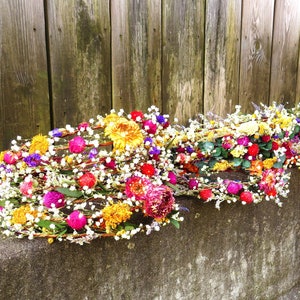May include: Several colorful floral crowns rest on a concrete ledge in front of a wooden fence. The crowns feature a variety of flowers in shades of yellow, pink, red, purple, and white. The crowns are made of woven branches.