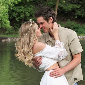 May include: A couple embraces near a lake. The woman wears a white off-the-shoulder top with lace sleeves and a matching skirt. She has a floral crown in her wavy blonde hair. The man wears a tan button-down shirt. The background features green trees and water.
