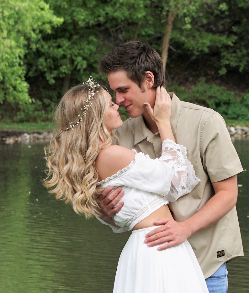 May include: A couple embraces near a lake. The woman wears a white off-the-shoulder top with lace sleeves and a matching skirt. She has a floral crown in her wavy blonde hair. The man wears a tan button-down shirt. The background features green trees and water.